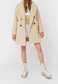 Eine Person trägt einen beigen Teddy-Mantel mit schwarzen Knöpfen, einen hellbraunen und weißen Kapuzenpullover sowie creamfarbene Schnürstiefel und steht vor einem weißen Hintergrund.