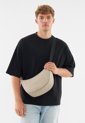 Beige leather crossbody bag with a curved design, featuring a smooth texture and an adjustable black strap. Simple logo detail on the front.