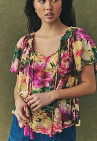 Bloem blouse met levendige roze, gele en groene patronen. Korte mouwen, een v-hals en strikjes met roze kwastjes voor detail.