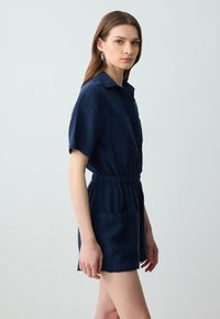 Combinaison en lin bleu marine avec des manches courtes, taille élastique et deux poches avant. Col simple et texture lisse, idéale pour les temps chauds.