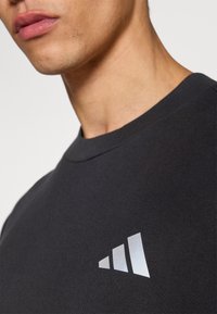 Černé bavlněné tričko s kulatým výstřihem, se reflexním logem se třemi pruhy na levé straně hrudi. Hladká textura a přiléhavý střih.