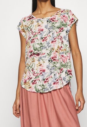 Femme portant un chemisier blanc à manches courtes avec un imprimé floral rose, vert et gris, associé à une jupe rose poussiére.