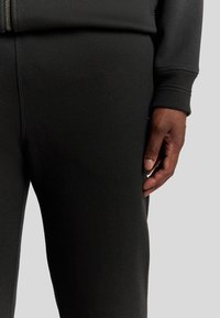 Schwarze, strukturierte Jogginghosen mit einem elastischen Bund, die eine Seitentasche und gerippte Bündchen haben. Die Hand ruht auf dem Stoff.
