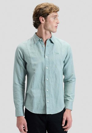 Junger Mann mit hellblauem Hemd mit Knöpfen, der nach rechts schaut, mit kurzem welligem Haar und sichtbarer schwarzer Hose.