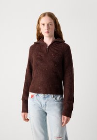 Bruine geribbelde trui met een ritskraag, gestructureerd patroon en kleurvlekken. Draagt bij lichtblauwe versleten jeans.