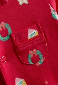Salopeta dintr-un material textil roșu, decorată cu imprimeuri tematice de sărbătoare, inclusiv coronițe și porumb fiert, având un detaliu mic de buzunar și închideri cu capse.