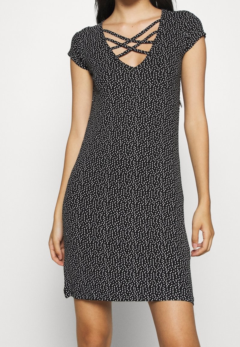 Femme portant une robe noire à manches courtes avec des pois blancs et des bretelles croisées au décolleté en V profond, debout devant un fond uni.