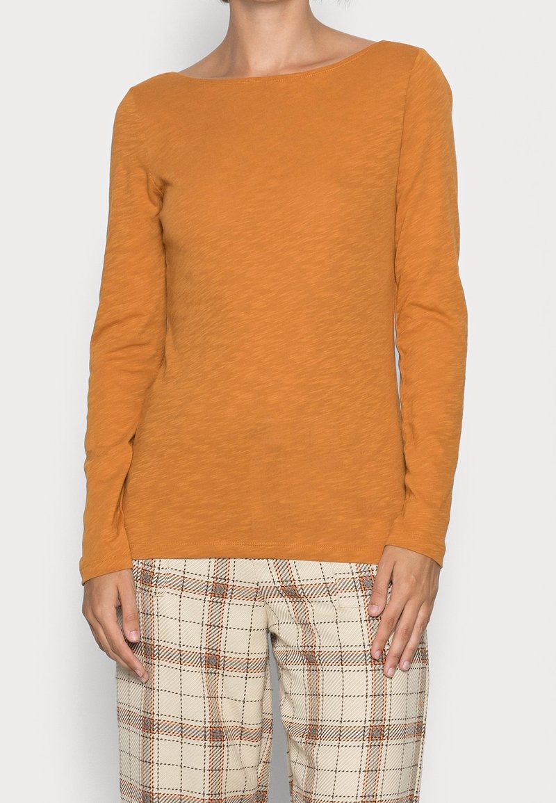 Maglietta a maniche lunghe di colore arancio bruciato, con scollo ampio e tessuto strutturato. Abbinata a pantaloni a quadri, beige chiaro e marrone.