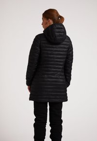 Giacca a vento nera con cappuccio, caratterizzata da sezioni orizzontali trapuntate, design lunghezza ginocchio e una texture liscia, indossata sopra pantaloni neri.