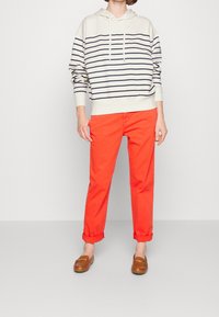 Gestreepte crèmekleurige hoodie met marineblauwe lijnen, gecombineerd met opgerolde, feloranje broeken en bruine instappers. Eenvoudig ontwerp en casual pasvorm.