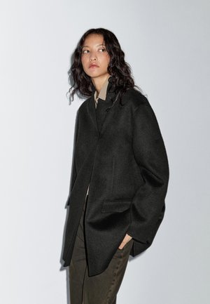 Schwarzer Wollmantel mit lockerer Passform, Reverskragen, Fronttaschen und minimalistischem Design. Weicher Stoff und mittellange Ärmel.