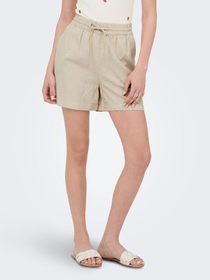 Kvinde iført beige linnedshorts med snor, hvid top med jordbærprint og beige slip-in sandaler, stående med den ene hånd hvilende på låret.