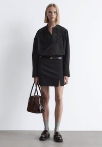 Woman standing against white wall wearing black embroidered blouse, black skirt with slit, gray socks, loafers, and holding brown leather handbag.
