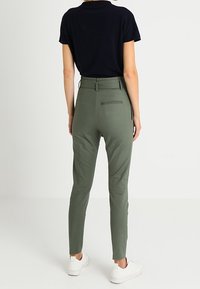 Gröna, åtsittande byxor med hög midja och bakfickor. Materialet verkar slätt, med en avsmalnande bendesign. Bärs med en marinblå topp och vita sneakers.