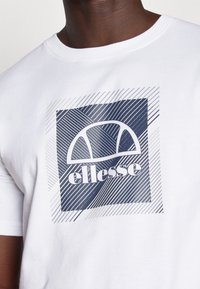 Camiseta de algodón blanca con un estampado gráfico en tonos azules con líneas diagonales y el logo de "ellesse" centrado dentro de un cuadrado.