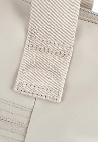 Beige tygväska rem med en texturerad ribbad design och sömnadsdetaljer. Remmen fästs vid en slät yta.