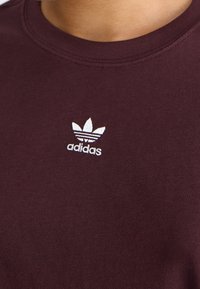 Burgundská bavlněná tričko s kulatým výstřihem a bílým vyšitým logem Adidas, které má tři pruhy ve tvaru listů. Jednoduchý, přiléhavý design.