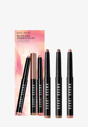 Set Bobbi Brown Golden Glow Shadow Stick montrant six ombres à paupières en stick noir dans des tons nude et bronze avec un fond rose et orange.