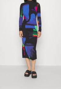 Femme portant une robe midi noire avec des motifs abstraits colorés et des sandales à plateforme noires, debout devant un mur blanc uni.