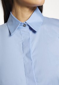 Light blue shirt featuring a large collar, button closure, and smooth texture with a subtle sheen. Details include a decorative button accent.