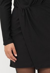 Abito nero con maniche lunghe, design arricciato in vita e orlo fluente. Il tessuto appare liscio con una leggera lucentezza.