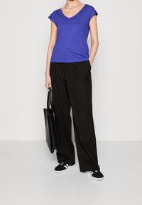 V-neck purple t-shirt, short sleeves, loose black trousers, black tote bag, and black sneakers with white soles in a plain background.