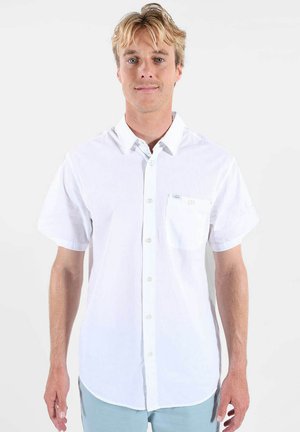 Homme blond portant une chemise blanche à manches courtes avec une poche poitrine, debout devant un fond clair uni.