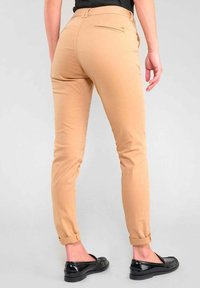 Person, der beige Slim-Fit-Hosen trägt, an den Knöcheln hochgerollt, und schwarze Slipper, steht vor einem schlichten hellen Hintergrund.