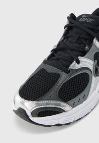 Buty sportowe Nike z czarną siateczkową cholewką, srebrnymi akcentami i fakturowanym wzorem. Posiadają białą podeszwę i czarne sznurowadła. Wytrzymała konstrukcja.