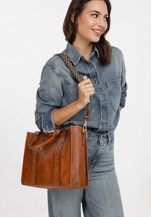 Vrouw in een spijkeroutfit draagt een grote bruine leren totebag met een schouderriem met patroon, ze glimlacht en kijkt naar rechts.