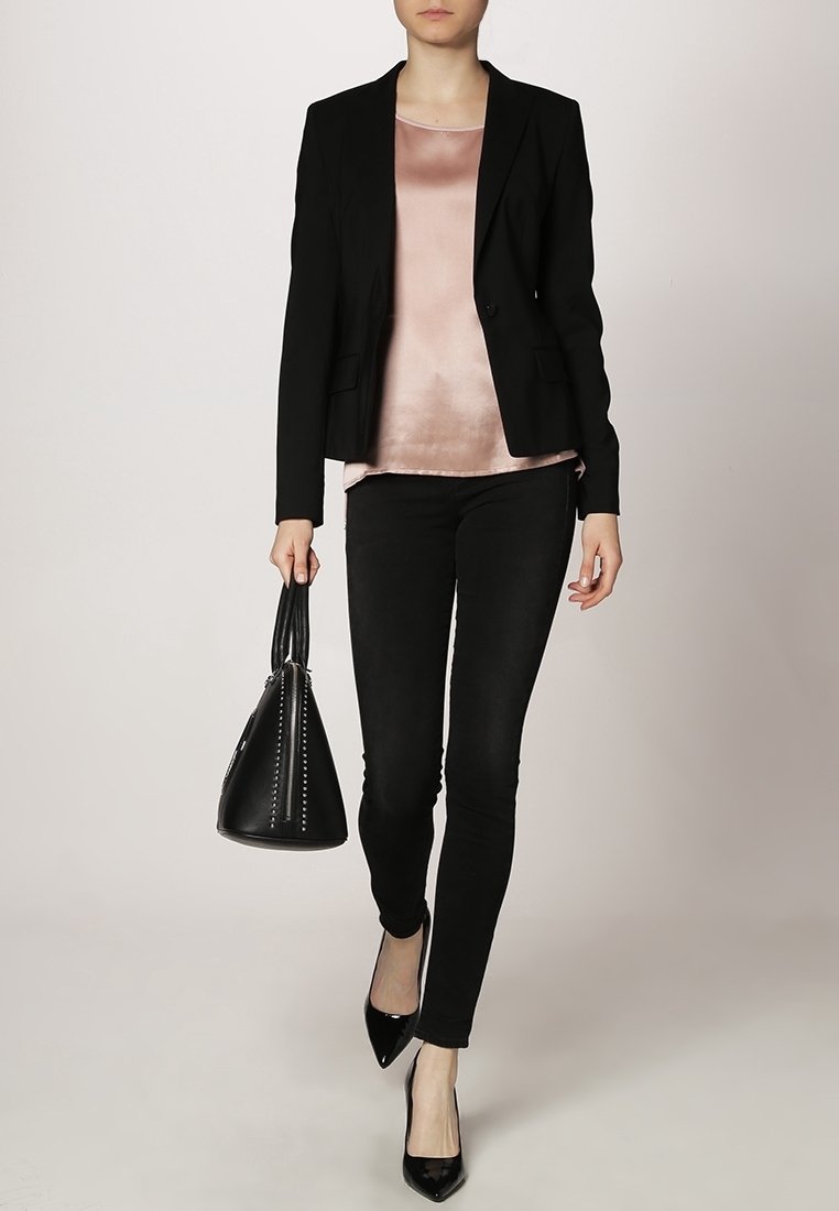 Femme portant un blazer noir, un haut en satin rose, un pantalon noir, des talons noirs, et tenant un sac à main noir.