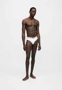 Modèle homme à la peau foncée portant un slip blanc de la marque Off-White, debout pieds nus sur un fond gris clair uni.