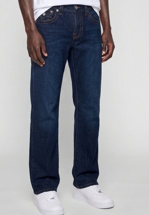 Mann trägt dunkelblaue Straight-Leg-Jeans und weiße Sneakers, steht vor einem einfarbigen hellen Hintergrund.