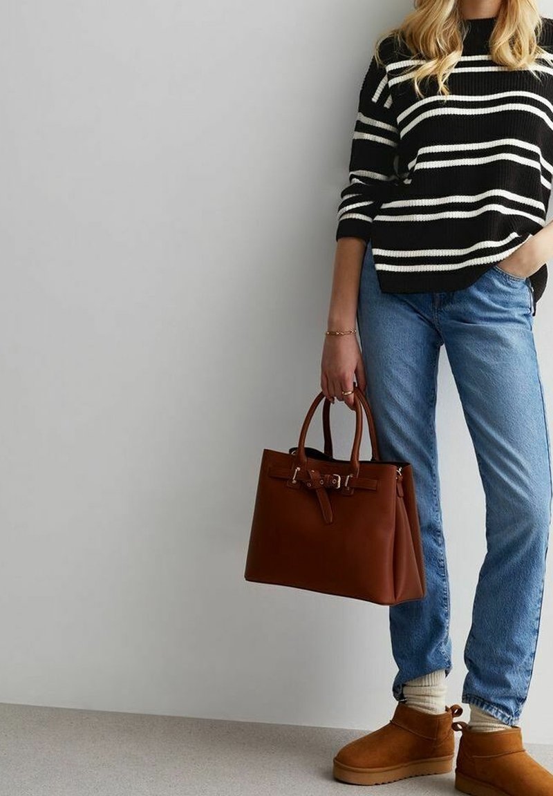 Borsa a mano in pelle marrone con un design strutturato, doppi manici e dettaglio con fibbia. Indossata con un maglione a righe bianche e nere e jeans blu.
