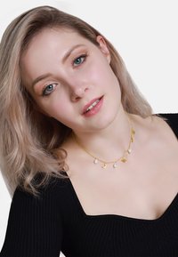 Collier en or avec des pendentifs en forme de lune croissante et d'étoile, orné de perles blanches. Le modèle porte un haut noir à côtes et a des cheveux blonds ondulés.