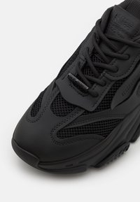 Zapatilla negra con parte superior de malla y sintético, con punta redondeada, cordones y acentos texturizados. Detalle del logo en la lengüeta.