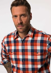 Camisa de franela a cuadros en rojo, azul marino y blanco, con cuello tipo botón, dos bolsillos en el pecho y botones negros. Textura suave y diseño clásico.