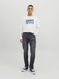 Camisa blanca de manga larga con texto gráfico azul, combinada con jeans oscuros ajustados y zapatillas negras con suelas blancas.
