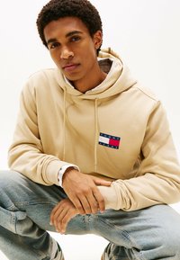 Beige hoodie med frontlomme, prydet med et rektangulært logo i rød, hvid og blå. Modellen bærer lyseblå denim jeans.