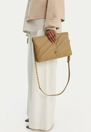 Persona con chaqueta beige y pantalones anchos blancos que sostiene un bolso acolchado color camel con cadena dorada a un lado.