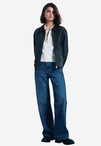 Grön kanvaskappa med brun krage, knäppning fram, sidofickor, kombinerad med högvikta blå denimjeans och svarta skor.