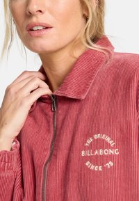 Giacca rossa in corduroy con zip e colletto, caratterizzata da un logo ricamato "BILLABONG" e una texture a coste. La zip e i polsini sono realizzati nello stesso materiale.
