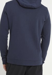 Felpa blu navy realizzata in tessuto morbido con tasca a canguro e polsini a coste. Presenta un cappuccio con cordini e un design dal fit rilassato.