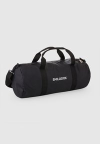 Musta duffel-laukku, joka on valmistettu kestävästä kankaasta. Siinä on kaksinkertaiset kahvat ja säädettävä olkahihna. Laukussa on vetoketjukiinnitys ja näkyvä valkoinen logo.