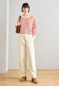 Randig stickad tröja i röd och kräm, beige byxor med hög midja och bruna klackstövlar. Håller en brun väska. Enkel, avslappnad look.