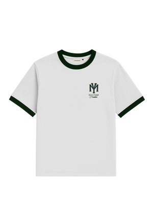 Giovane uomo con t-shirt bianca con bordatura verde, sciarpa al collo verde scuro a fantasia, pantaloni beige e cappellino bianco con logo blu scuro.