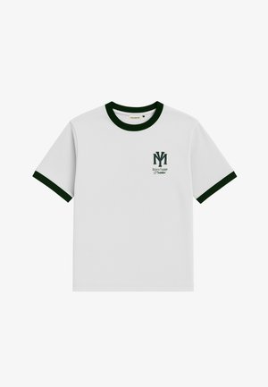 Giovane uomo con t-shirt bianca con bordatura verde, sciarpa al collo verde scuro a fantasia, pantaloni beige e cappellino bianco con logo blu scuro.