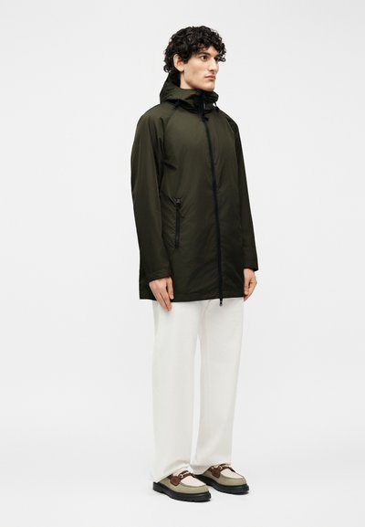 Jeune homme debout portant une veste à capuche vert foncé, un pantalon blanc et des chaussures beige et marron sur un fond clair uni.