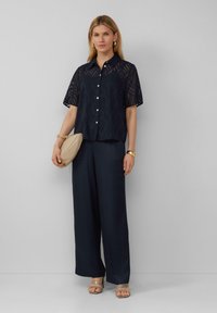 Marineblauw shirt met korte mouwen en golfpatroon, knoopsluiting aan de voorkant, gecombineerd met marineblauwe wijde broek en tan blokhak sandalen. Beige clutch tas.