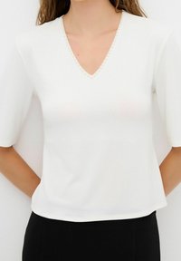 Blusa blanca de manga corta con escote en V, que presenta una fila de pequeñas perlas a lo largo del borde. Tejido suave con una textura lisa.